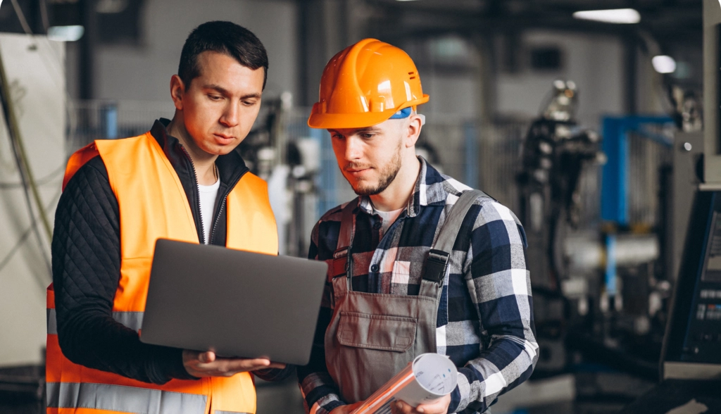 Workers in safety gear reviewing equipment data on a laptop in an industrial facility, representing standardized asset information.